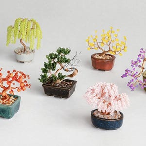 May include: Six miniature bonsai trees crafted from wire and colourful beads, each planted in a small ceramic pot filled with pebbles. The trees feature various colours including light green, orange, dark green, yellow, pink, and purple.