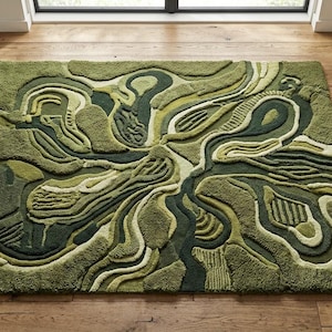 Op de afbeelding: Een groot, rechthoekig groen tapijt met een abstract ontwerp. Het tapijt heeft een gelaagd patroon in groentinten, waardoor een gestructureerd, driedimensionaal effect ontstaat. Het tapijt ligt op een houten vloer, met een bank en een bijzettafel.