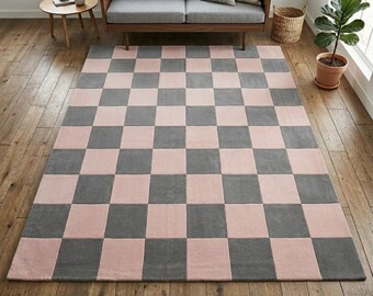 May include: A large rectangular rug featuring a checkerboard design of grey and pink squares. The rug is laid on a wooden floor, creating a neutral and modern aesthetic. This rug is a home decor item, suitable for various living spaces.