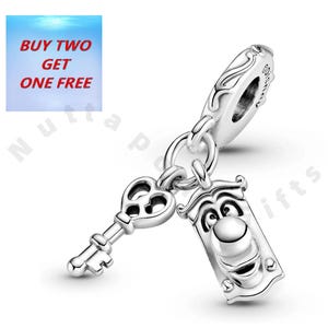 Może przedstawiać: Srebrny charms przedstawiający klucz i kołatkę do drzwi z Alicji w Krainie Czarów. Kołatka ma uśmiechniętą twarz. Charms jest przymocowany do srebrnej pętli. Tekst "BUY TWO GET ONE FREE" znajduje się w lewym górnym rogu.