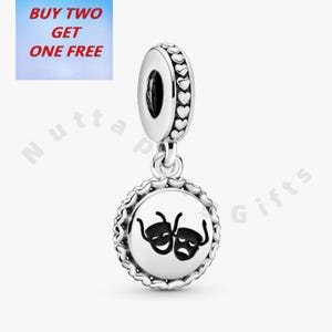 Op de afbeelding: Een zilveren bedel met een theaterthema. De bedel heeft een ronde hanger met twee zwarte theatermaskers. De bovenkant van de bedel heeft een hartvormig ontwerp. De afbeelding bevat ook de tekst "BUY TWO GET ONE FREE".