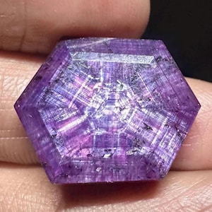 Puede incluir: Una gema hexagonal facetada de color púrpura. La gema tiene un tono violeta intenso con líneas más claras e intersecantes que crean un patrón único. Pequeñas inclusiones oscuras son visibles en toda la piedra, lo que aumenta su belleza natural.