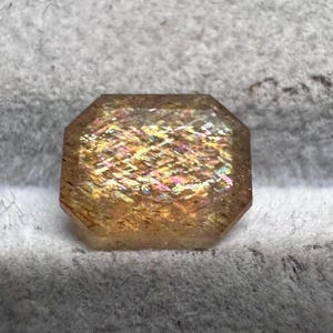 May include: A faceted, rectangular gemstone with a warm, golden-brown hue. The gem displays internal iridescence with flashes of yellow, pink, and orange. The stone is set against a soft, gray background.