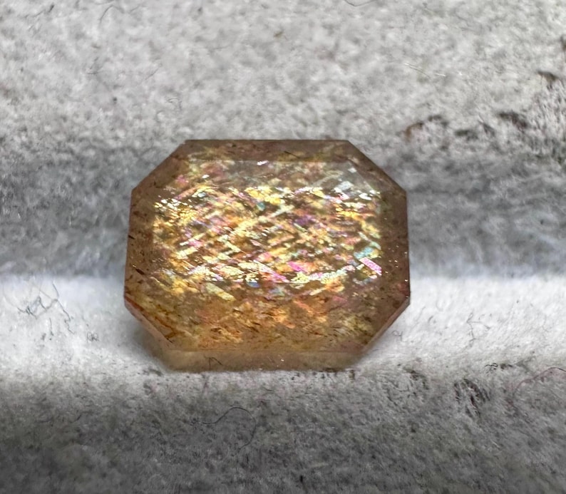 May include: A faceted, rectangular gemstone with a warm, golden-brown hue. The gem displays internal iridescence with flashes of yellow, pink, and orange. The stone is set against a soft, gray background.