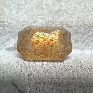 May include: A faceted, rectangular gemstone with a warm, golden-orange hue. The gem has a textured appearance and displays internal color flashes. The stone is set against a soft, white background.