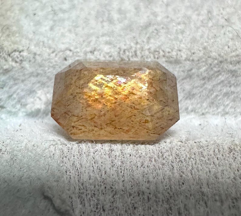 May include: A faceted, rectangular gemstone with a warm, golden-orange hue. The gem has a textured appearance and displays internal color flashes. The stone is set against a soft, white background.