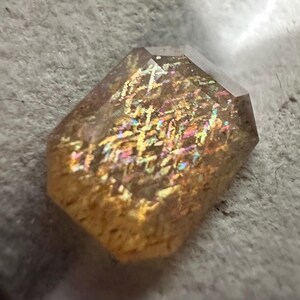 May include: A faceted, rectangular gemstone with a warm, golden-brown hue. The stone displays internal iridescence with flashes of rainbow colors, including pink, blue, and yellow. The gem is set against a textured, light gray surface.