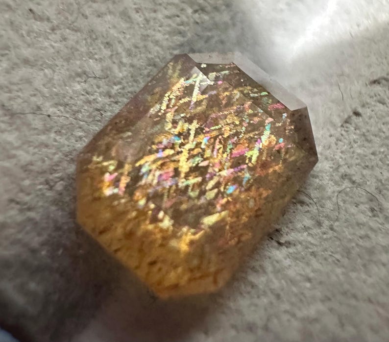 May include: A faceted, rectangular gemstone with a warm, golden-brown hue. The stone displays internal iridescence with flashes of rainbow colors, including pink, blue, and yellow. The gem is set against a textured, light gray surface.