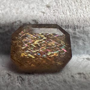 May include: An octagonal-cut gemstone with a warm, golden-brown hue. The stone displays a shimmering, iridescent effect with flashes of yellow, pink, and blue. The gemstone is set against a soft, textured background.