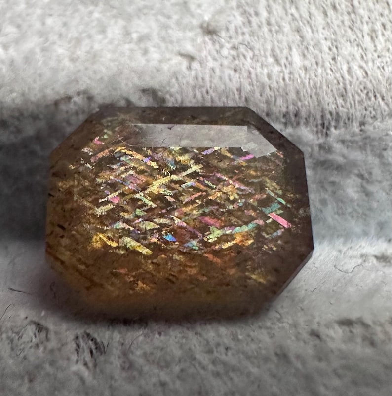 May include: An octagonal-cut gemstone with a warm, golden-brown hue. The stone displays a shimmering, iridescent effect with flashes of yellow, pink, and blue. The gemstone is set against a soft, textured background.