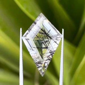 以下が含まれることがあります： 黒い針状のインクルージョンが入った、透明な菱形の宝石。銀色のピンセットで保持されています。宝石はカットされており、光を捉え、内部の模様を表示します。背景は柔らかな緑色です。