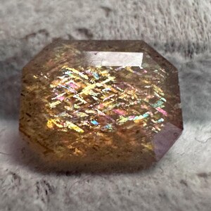 May include: A faceted gemstone with a rectangular shape and beveled edges. The stone has a warm, brown base color with internal flashes of iridescent colors, including yellow, pink, and blue. The gem is cut to maximize light reflection.
