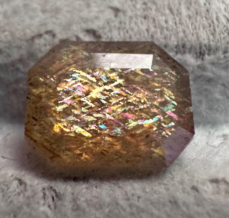 May include: A faceted gemstone with a rectangular shape and beveled edges. The stone has a warm, brown base color with internal flashes of iridescent colors, including yellow, pink, and blue. The gem is cut to maximize light reflection.