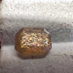 May include: A faceted gemstone with a rectangular shape and beveled edges. The gem has a golden-brown hue with iridescent flashes of pink, yellow, and green. The stone is set against a soft, gray background.