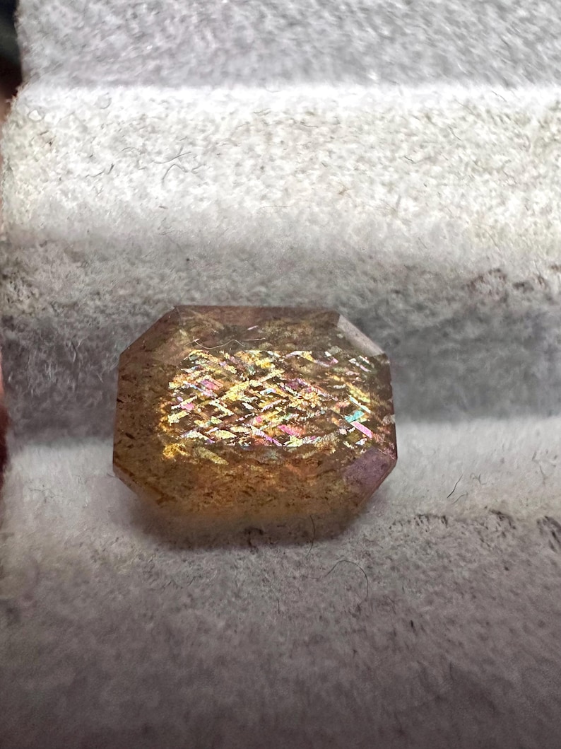 May include: A faceted gemstone with a rectangular shape and beveled edges. The gem has a golden-brown hue with iridescent flashes of pink, yellow, and green. The stone is set against a soft, gray background.