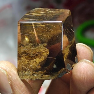 May include: A transparent, faceted crystal with golden, needle-like inclusions. The crystal has a warm, amber hue and a rough, textured base. The interior features a network of golden strands and a small, golden sphere.