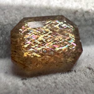 May include: A faceted gemstone with a warm, golden-brown base color. The stone displays iridescent flashes of pink, yellow, and blue across its surface. The cut is an octagon shape, with a polished, reflective finish.