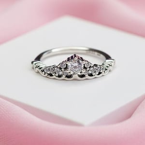 May include: A silver-colored ring with a crown design. The ring features a central, round, light-colored stone and smaller stones along the crown's base. The ring sits on a white surface against a pink background.