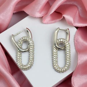May include: A pair of silver-coloured link earrings. Each earring has a large, oval-shaped link encrusted with small, sparkling crystals. The earrings are displayed on a white surface with a pink satin backdrop.