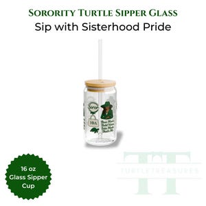 Op de afbeelding: Een doorzichtige glazen sipperbeker met een bamboe deksel en rietje. De beker heeft groene en witte afbeeldingen, waaronder de tekst "Sorority Turtle Sipper Glass" en "Sip with Sisterhood Pride". De beker bevat 473 ml.