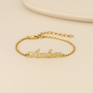 Peut inclure: Un bracelet doré avec le nom "Amber" écrit en cursive. Le bracelet est composé d'une chaîne délicate et d'un fermoir. Le bijou est présenté sur une surface de couleur claire.