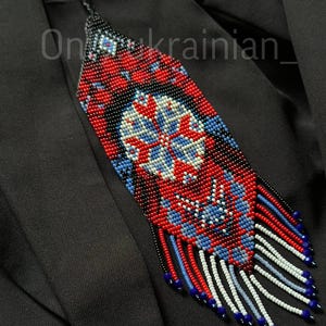 May include: A beaded pendant necklace with a geometric design in red, blue, white, and black. The pendant has a central floral motif and a fringe of beads at the bottom. The text "On_ukrainian_" is visible on the image.