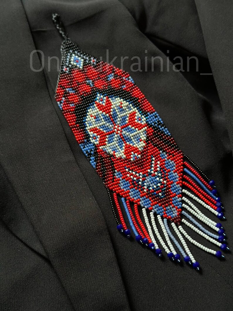 May include: A beaded pendant necklace with a geometric design in red, blue, white, and black. The pendant has a central floral motif and a fringe of beads at the bottom. The text "On_ukrainian_" is visible on the image.