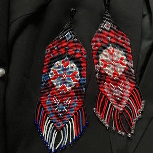 Könnte beinhalten: Zwei Ohrringe mit Perlenbesatz und aufwendigen Mustern. Die Ohrringe weisen eine Kombination aus roten, blauen, weißen und schwarzen Perlen auf. Das Design umfasst florale und geometrische Motive mit hängenden Perlenfransen.