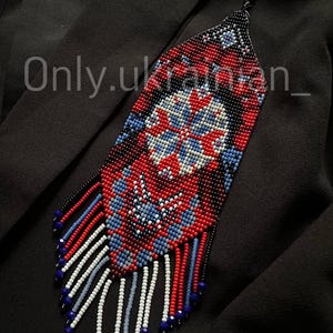 May include: A beaded necklace with a geometric design in red, white, and blue. The necklace has a long, rectangular shape with a fringe of beads at the bottom. The text "Only.ukrainian_" is visible on the left side.