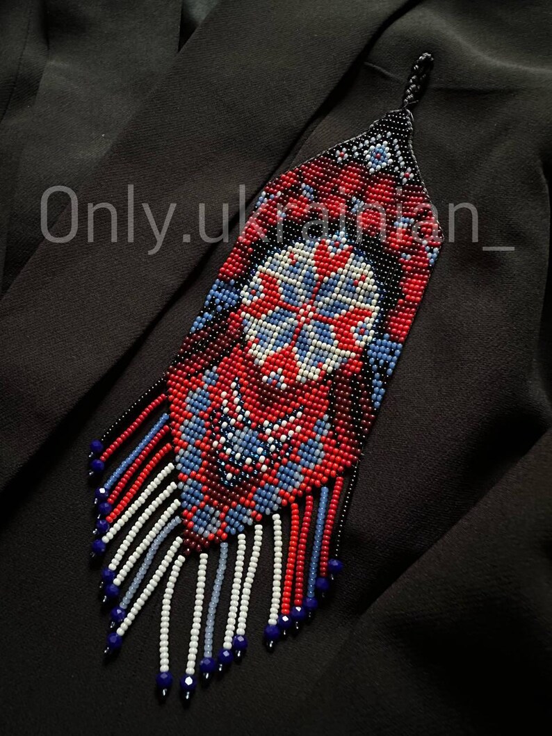 May include: A beaded necklace with a geometric design in red, white, and blue. The necklace has a long, rectangular shape with a fringe of beads at the bottom. The text "Only.ukrainian_" is visible on the left side.