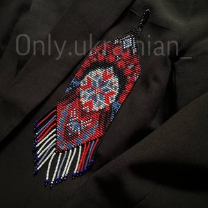 May include: A beaded pendant with a geometric design in red, blue, white, and black. The pendant has a long, fringed bottom and is displayed against a black background. The text "Only.ukrainian_" is visible.