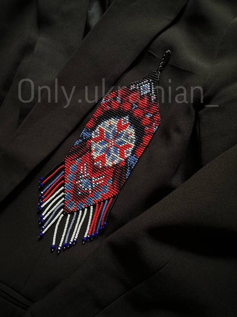 May include: A beaded pendant with a geometric design in red, blue, white, and black. The pendant has a long, fringed bottom and is displayed against a black background. The text "Only.ukrainian_" is visible.