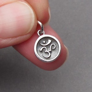 May include: A small, round silver pendant featuring the Om symbol. The pendant has a detailed, raised design of the sacred syllable within a circular frame. The pendant is attached to a small loop for a chain.