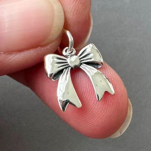 May include: A small, silver-colored bow charm with a loop for hanging. The bow has detailed lines and a polished finish, with the ends of the ribbon curving downwards. The charm is held between two fingers.