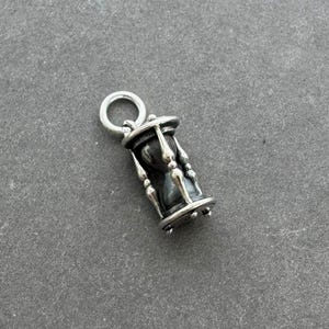 May include: A silver hourglass charm with a black sand-filled glass. The charm has a loop at the top for attaching to a necklace or bracelet.