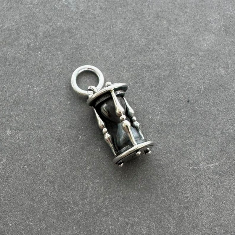 May include: A silver hourglass charm with a black sand-filled glass. The charm has a loop at the top for attaching to a necklace or bracelet.