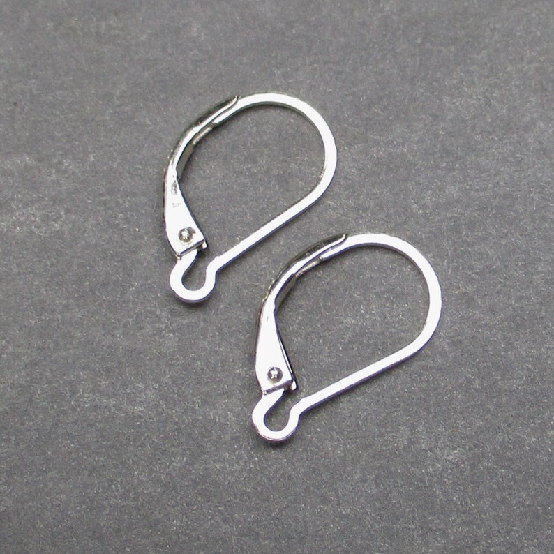 May include: Two silver-colored leverback earring findings. The findings are made of metal and have a curved, looped design with a hinged lever closure. The background is a solid, dark gray color.