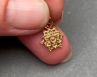 Op de afbeelding: Een kleine goudkleurige metalen bedel met een symmetrisch, meerlaags bloem- of mandala-ontwerp met een centrale cirkelvormige stip en puntige bloemblaadjes die naar buiten stralen, compleet met een klein ringetje aan de bovenkant.