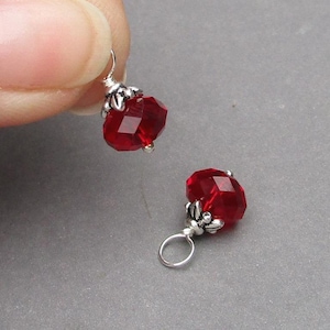May include: Two faceted, red crystal earrings with silver-coloured metal accents. One earring is held by a hand, while the other rests on a grey surface. The crystals have a geometric shape.