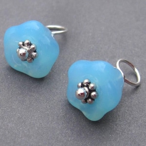 May include: Two light blue flower-shaped earrings with silver-colored accents. Each earring has a small, round, silver-colored center and a silver loop for hanging. The earrings are set against a gray background.