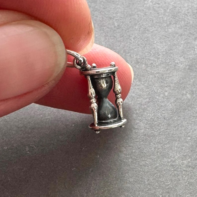 May include: A silver charm in the shape of an hourglass. The hourglass is detailed with a black sand effect and ornate silver accents.