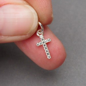 May include: A small silver cross pendant featuring a series of clear, sparkling stones embedded along the front surface. The cross includes a small circular jump ring at the top for attachment, showcasing a metallic finish and reflective accents.