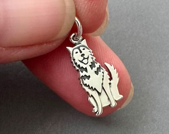 May include: A small silver-toned metal charm shaped like a sitting husky dog. The charm features detailed cutouts and engraved lines to define the dog's facial features, fur texture, and upright ears, with a small circular jump ring at the top.