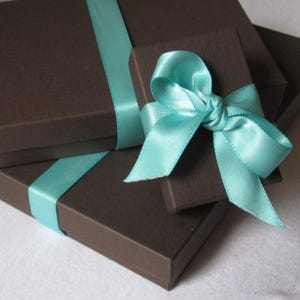 K&ouml;nnte beinhalten: Zwei rechteckige, dunkelbraune Geschenkboxen mit strukturierter Oberfl&auml;che. Die gr&ouml;&szlig;ere Box ist mit einem glatten, hellblauen Satinband umwickelt. Auf ihr liegt eine kleinere Box, die mit einer hellblauen Satinschleife verziert ist.