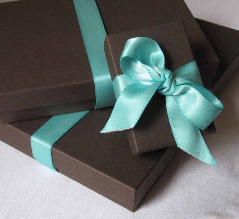 K&ouml;nnte beinhalten: Zwei rechteckige, dunkelbraune Geschenkboxen mit strukturierter Oberfl&auml;che. Die gr&ouml;&szlig;ere Box ist mit einem glatten, hellblauen Satinband umwickelt. Auf ihr liegt eine kleinere Box, die mit einer hellblauen Satinschleife verziert ist.