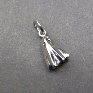 May include: A polished silver-toned metal charm shaped like a ghost with two circular cutouts for eyes and a draped, flowing body. The charm includes a small circular jump ring attached to the top, allowing it to be suspended from a chain or bracelet.
