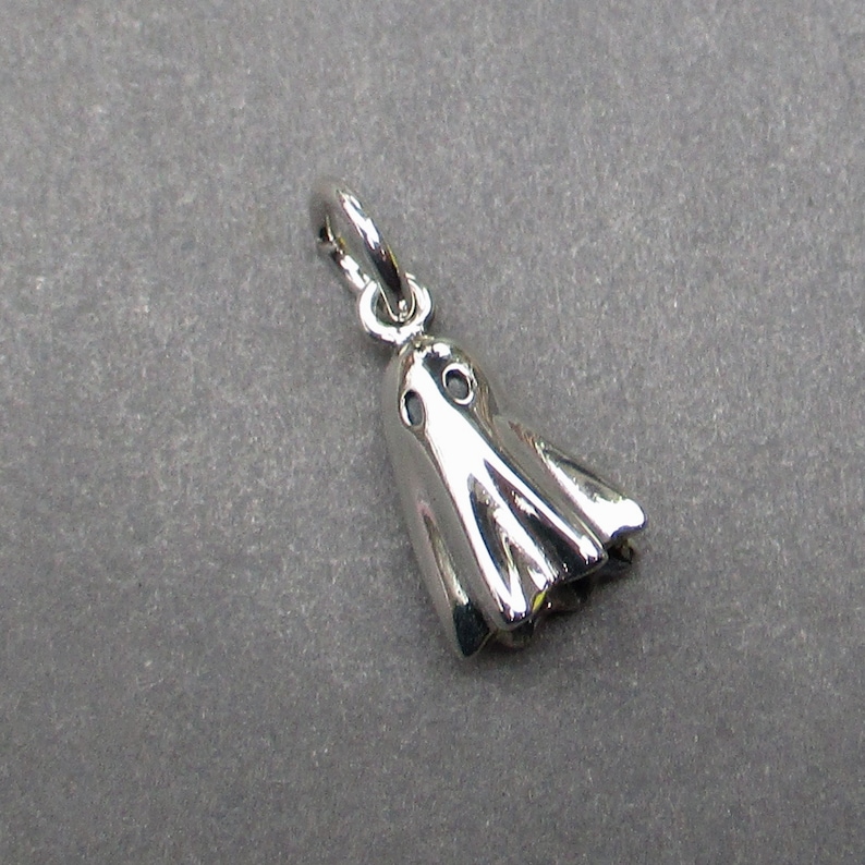 May include: A polished silver-toned metal charm shaped like a ghost with two circular cutouts for eyes and a draped, flowing body. The charm includes a small circular jump ring attached to the top, allowing it to be suspended from a chain or bracelet.