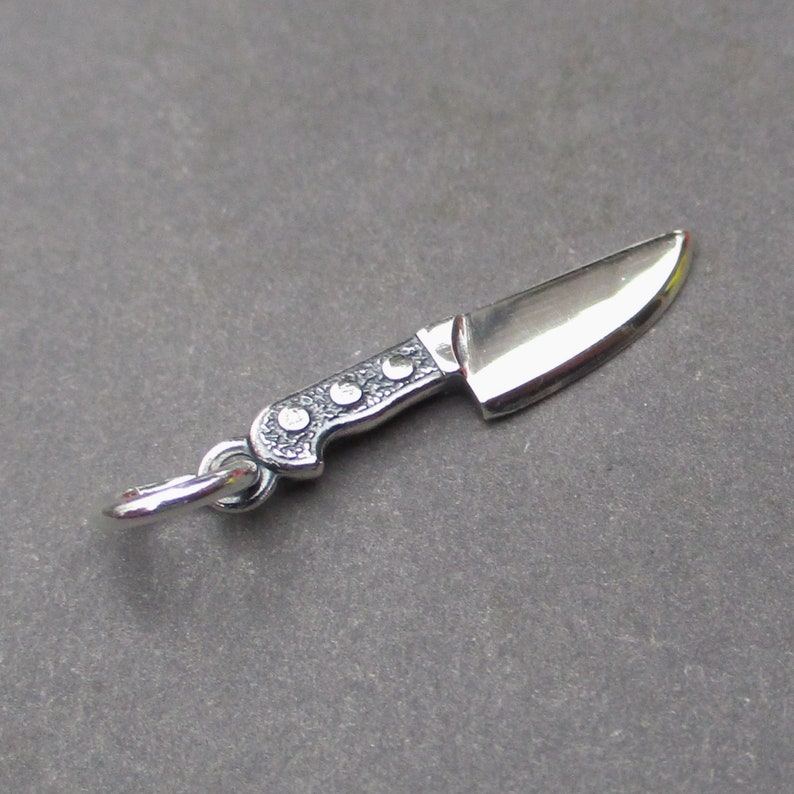 May include: A silver knife charm with a detailed handle and a sharp blade. The charm has a loop for attaching to a necklace or bracelet.