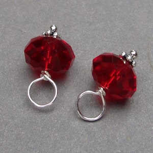 May include: Two faceted red crystal beads, each attached to a silver-toned metal jump ring. The beads are topped with small, decorative silver-toned metal accents. These components are designed for jewellery making and crafting projects.