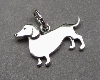 May include: A polished silver-toned metal charm shaped like a dachshund dog in profile, featuring a smooth, reflective surface and a circular jump ring attached to the top for hanging. The charm depicts the dog standing with its tail slightly raised.